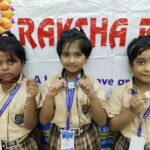 Three young students in beige uniforms with blue lanyards hold crafts at a school event in front of a colorful banner